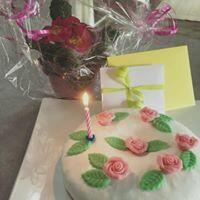 Ein weißer Kuchen mit rosa Zuckerguss und grünen Blättern liegt auf einem Teller. Eine einzelne brennende Kerze befindet sich obenauf. Hinter der Torte stehen eine Topfpflanze, ein gelber Umschlag und eine kleine verpackte Karte.