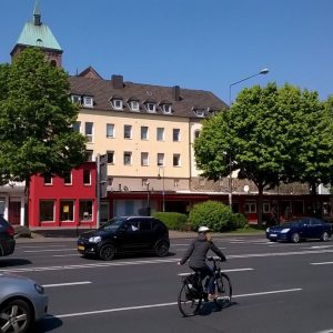 Ein Mann fährt mit dem Fahrrad auf einer Stadtstraße mit vorbeifahrenden Autos. Im Hintergrund befindet sich ein mehrstöckiges beigefarbenes Gebäude mit einem grünen Turmdach, umgeben von Bäumen und einem roten Schaufenster auf Straßenniveau.