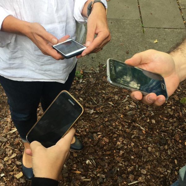 Drei Personen stehen im Freien in einem Kreis, jede hält ein Smartphone in der Hand und schaut darauf. Der Boden ist mit Holzspänen und Pflastersteinen bedeckt, und nur ihre Hände und Oberkörper sind sichtbar.