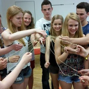 Zehn Teenager stehen im Kreis, lächeln und halten miteinander verbundene Schnüre, die in einem Klassenzimmer ein Netz bilden.