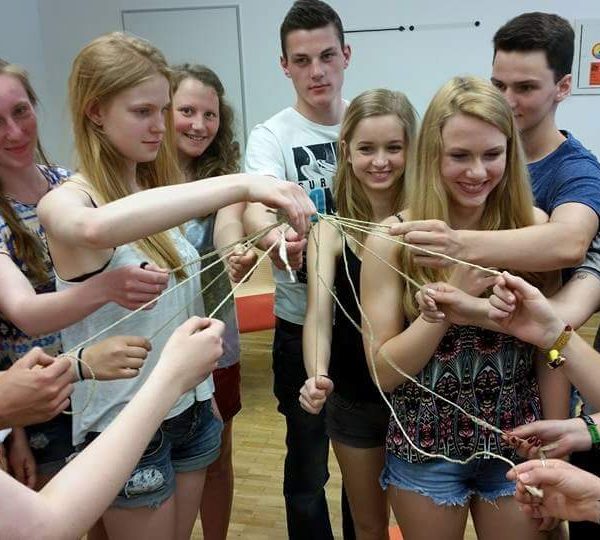 Zehn Teenager stehen im Kreis, lächeln und halten miteinander verbundene Schnüre, die in einem Klassenzimmer ein Netz bilden.