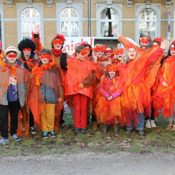 Eine Gruppe von Menschen in leuchtend roten und orangefarbenen Kostümen mit Masken und Umhängen steht im Freien vor einem Gebäude, lächelt und winkt enthusiastisch in die Kamera.