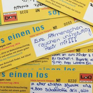 Mehrere gelbe Karten mit handgeschriebenen deutschen Botschaften über die Gefahren des Rauchens liegen verstreut auf einem Tisch. Die wichtigste sichtbare Karte warnt vor den Folgen des Passivrauchens für die Angehörigen.