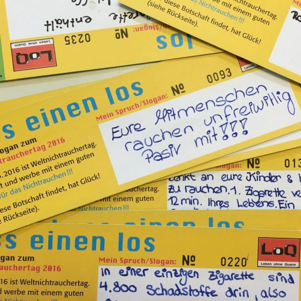 Mehrere gelbe Karten mit handgeschriebenen deutschen Botschaften über die Gefahren des Rauchens liegen verstreut auf einem Tisch. Die wichtigste sichtbare Karte warnt vor den Folgen des Passivrauchens für die Angehörigen.