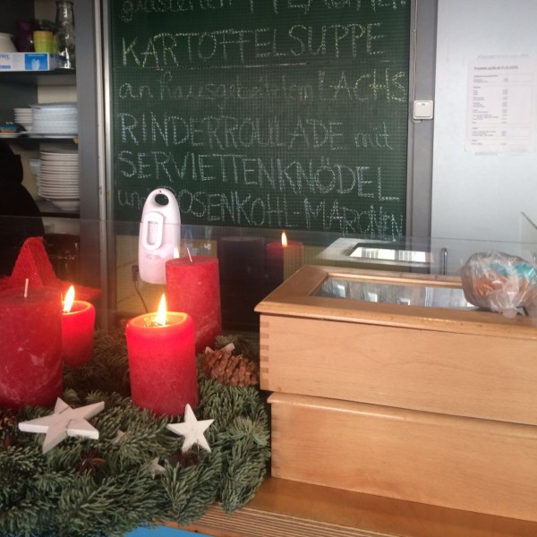 Über einem Tresen mit Kerzen, Tannenzweigen und Holzsternen hängt eine deutschsprachige Speisekarte mit Tagesangeboten. Auf der Karte stehen Entenbrust, Kartoffelsuppe, Rinderroulade, Knödel und Apfelmus.