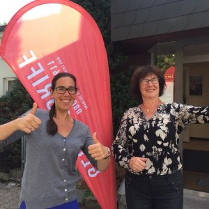 Zwei lächelnde Frauen stehen draußen in der Nähe eines roten Fahnenbanners. Beide zeigen den Daumen nach oben; die Frau links trägt eine Brille und einen grauen Pullover, die Frau rechts trägt eine Brille und ein schwarzes geblümtes Oberteil. Sie stehen vor einem Gebäudeeingang.