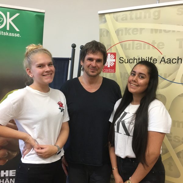 Drei Personen stehen lächelnd vor Bannern der AOK, der Sucht Hilfe Aachen und der Caritas. Jeweils zwei junge Frauen stehen neben einem Mann in der Mitte, alle sind leger gekleidet und wirken glücklich.