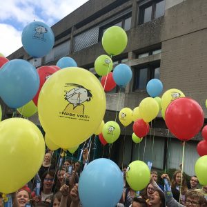 Eine Gruppe lächelnder Kinder im Freien hält bunte rote, gelbe und blaue Luftballons vor einem grauen Gebäude. Die gelben Luftballons sind mit einem Cartoon-Gesicht und deutschem Text bedruckt. Der Himmel ist teilweise bewölkt.
