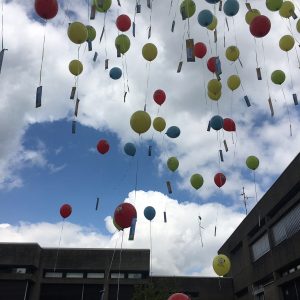 Bunte Luftballons mit angehängten Anhängern schweben vor einem teilweise bewölkten Himmel in die Höhe, eingerahmt von den dunklen Kanten zweier moderner Gebäude.