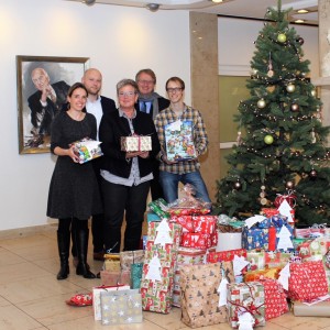 Fünf Personen stehen lächelnd vor einem geschmückten Weihnachtsbaum in einem Innenraum, umgeben von einem großen Stapel eingepackter Geschenke. Im Hintergrund sind ein Porträt und eine weiße Büste zu sehen.