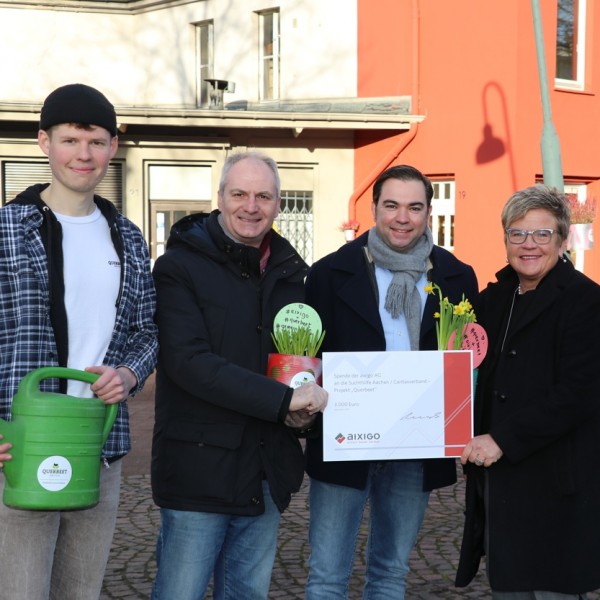 Vier Personen stehen lächelnd im Freien und halten eine große Urkunde, Blumen und eine Gießkanne in der Hand. Sie sind warm gekleidet, mit bunten Gebäuden und ein paar Fußgängern im Hintergrund.