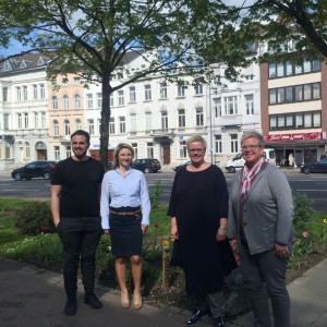 Vier Personen stehen lächelnd vor einem Garten mit Bäumen und einer von hohen historischen Gebäuden gesäumten Stadtstraße im Hintergrund. Es ist ein sonniger Tag.