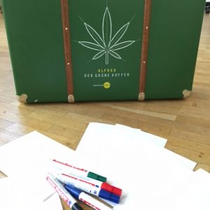 Ein grüner Koffer mit einem großen, mit Linien gezeichneten Cannabisblatt und dem Text Alfred der Grüne Koffer steht auf einem Holzboden, umgeben von verstreuten weißen Papieren und farbigen Stiften.