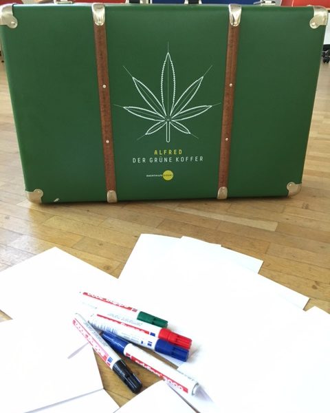 Ein grüner Koffer mit einem großen, mit Linien gezeichneten Cannabisblatt und dem Text Alfred der Grüne Koffer steht auf einem Holzboden, umgeben von verstreuten weißen Papieren und farbigen Stiften.