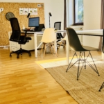 Ein helles, modernes Büro mit Holzböden, weißen Schreibtischen, schwarzen und weißen Stühlen, einer Korkwand mit Notizen, einer Uhr und großen Fenstern, die natürliches Licht hereinlassen. Auf dem Boden im Vordergrund liegt ein gewebter Teppich.
