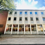 Ein dreistöckiges Gebäude mit großen Fenstern im Erdgeschoss, durch die eine rote Schrift zu sehen ist. Die Fassade ist weiß und aus Ziegeln, mit Bäumen und einem blauen Himmel darüber.