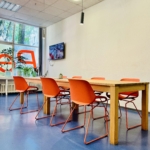 Ein moderner Besprechungsraum mit einem Holztisch, umgeben von sechs leuchtend orangefarbenen Stühlen auf einem blauen Boden. An der Wand ist ein Fernseher angebracht, und große Fenster lassen natürliches Licht herein.