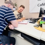 Zwei Männer sitzen sich an einem Büroschreibtisch gegenüber, lächeln und sehen sich gemeinsam Dokumente an. Auf dem Schreibtisch stehen Büromaterialien und ein Computermonitor, und an der Wand hinter ihnen ist ein Schild teilweise sichtbar.