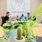 Zwei Personen arbeiten an Computern in einem Büro mit einem großen Café-Plattform-Schild mit Handsymbolen an der Wand; mehrere Topfpflanzen, darunter ein Kaktus, sind im Vordergrund zu sehen.
