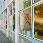 Die Fenster eines Gebäudes sind mit roten Handlinien auf Milchglas verziert. Im Inneren sind Grünpflanzen zu sehen, und das Sonnenlicht spiegelt sich auf dem Gehweg und den Fenstern. Draußen sind ein Baum und eine Straßenszene zu sehen.