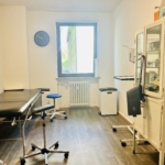 Ein sauberer, gut beleuchteter medizinischer Untersuchungsraum mit einem schwarzen Untersuchungstisch, einem blauen Hocker, weißen Schränken mit medizinischem Material, einer Uhr an der Wand und einem Fenster, das Tageslicht hereinlässt.
