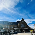 Ein großer steinerner Bahnhof mit einer Uhr über dem Eingang und einem "DB Hauptbahnhof"-Schild. Davor gehen Menschen, und links ist eine Pferdestatue zu sehen. Der Himmel über uns ist strahlend blau mit vereinzelten Wolken.