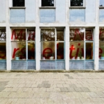 Auf den Fenstern des Erdgeschosses eines modernen Gebäudes steht in großen roten Buchstaben "Mit Nahe(r) Perspektive" auf Milchglas, und im Inneren sind Pflanzen zu sehen. Der Bürgersteig vor dem Gebäude ist leer.