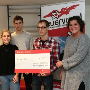 Fünf Personen stehen in einem Raum und halten einen großen rot-weißen Spendenscheck über 2.000 Euro in der Hand, im Hintergrund ein Banner des Feuervogels Aachen. Alle lächeln und schauen in die Kamera.