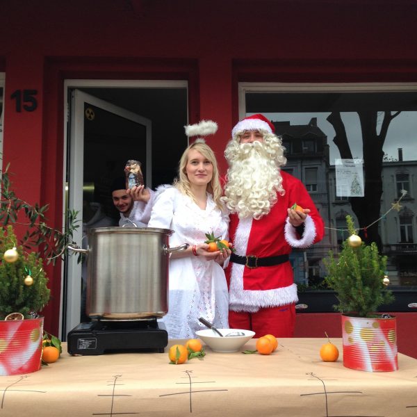 Eine als Engel verkleidete Frau und ein als Weihnachtsmann verkleideter Mann stehen lächelnd vor einem roten Gebäude. Auf einem Tisch steht ein großer Topf, der mit Orangen, Grünzeug und Ornamenten geschmückt ist. Eine weitere Person steht in der Tür hinter ihnen.