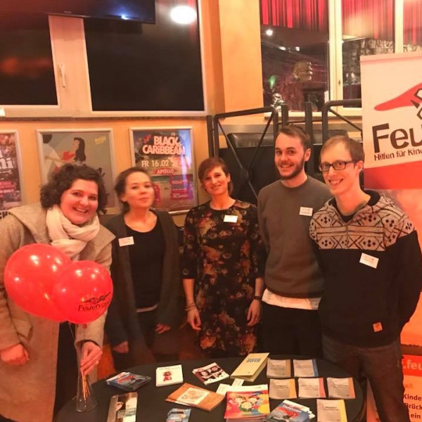 Fünf Personen stehen lächelnd hinter einem Tisch mit Broschüren und Luftballons bei einer Indoor-Veranstaltung. Ein Banner mit der Aufschrift "Feuervogel" und einem roten Vogellogo wirbt für eine Hilfsorganisation für Kinder suchtkranker Eltern.