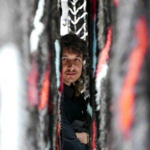 Eine Person mit langen Haaren und Schnurrbart blickt mit konzentriertem Gesichtsausdruck durch einen schmalen Spalt zwischen strukturierten, mehrfarbigen Stoffbahnen.