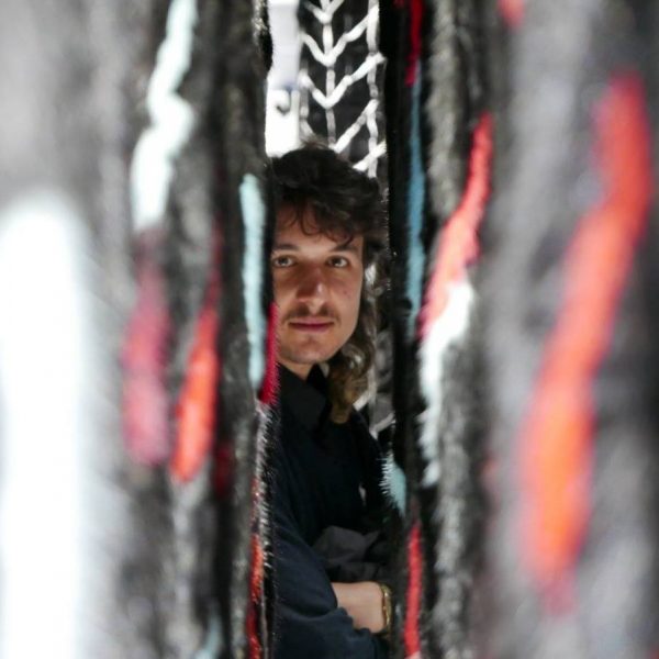 Eine Person mit langen Haaren und Schnurrbart blickt mit konzentriertem Gesichtsausdruck durch einen schmalen Spalt zwischen strukturierten, mehrfarbigen Stoffbahnen.