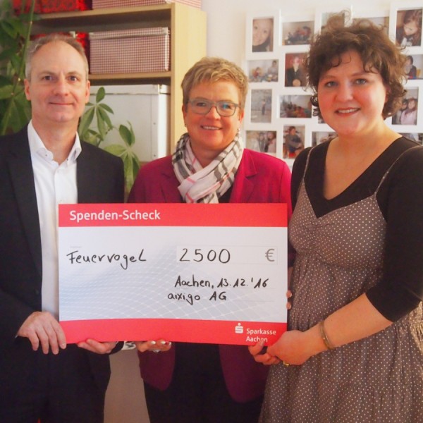 Drei Personen stehen drinnen und halten einen großen Spendenscheck über 2500 Euro, ausgestellt auf Feuervogel. Sie lächeln, und im Hintergrund sind Fotos und eine Pflanze zu sehen.