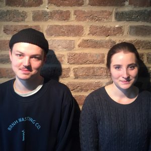 Zwei junge Erwachsene, ein Mann und eine Frau, stehen nebeneinander an einer Backsteinmauer und blicken beide in die Kamera. Der Mann trägt eine schwarze Mütze und ein dunkles Sweatshirt, die Frau hat ihr braunes Haar zurückgebunden und trägt einen dunklen Pullover.