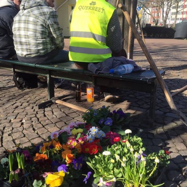 Eine Person, die eine gelbe Querbeet-Weste trägt, sitzt auf einer Bank neben zwei anderen Personen auf einem Kopfsteinpflaster, während in der Nähe bunte Topfblumen und Gartengeräte auf dem Boden liegen.