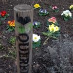 Ein Holzpfosten mit der Aufschrift LOVE OUR steht in einem Gartenbeet, in dem verschiedene bunte Blumen in der Erde blühen. Auch etwas Grünzeug und kleine Sträucher sind zu sehen.