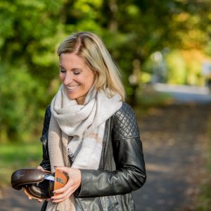 Eine lächelnde Frau mit blondem Haar, die einen Schal und eine schwarze Lederjacke trägt, steht auf einem von Bäumen gesäumten Weg und betrachtet eine kleine braune Brieftasche.