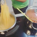 Eine Person in einem grünen Hemd kocht in einer Küche und hält trockene Spaghetti über einen Topf mit kochendem Wasser. Daneben steht ein Topf mit Tomatensoße, auf dem ein gelber Löffel ruht.