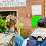 Zwei Personen mit Rucksäcken stehen vor einem Backsteingebäude und blicken auf ein Schild mit der Aufschrift "Café Plattform". Eine hat Haarspangen und telefoniert, die andere trägt einen Kapuzenpulli. Im Hintergrund ist üppiges Grün zu sehen.