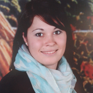 Eine junge Frau mit glattem dunklem Haar und heller Haut lächelt in die Kamera. Sie trägt einen hellblauen und weißen Schal und ein dunkles Oberteil. Der Hintergrund im Freien ist unscharf.