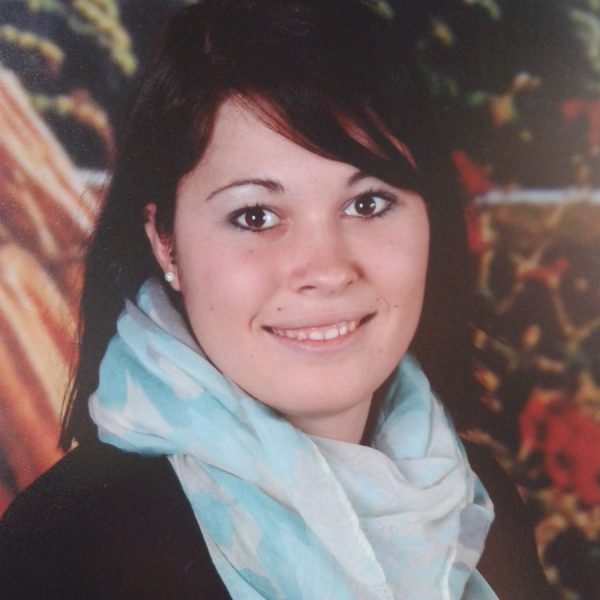 Eine junge Frau mit glattem dunklem Haar und heller Haut lächelt in die Kamera. Sie trägt einen hellblauen und weißen Schal und ein dunkles Oberteil. Der Hintergrund im Freien ist unscharf.