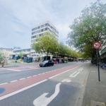 Eine Stadtstraße mit einem Tempo 30-Schild, Fahrradspuren und entlang des Bordsteins geparkten Autos. Bäume säumen den Gehweg, und ein mittelhohes Bürogebäude ist unter dem bewölkten Himmel zu sehen.