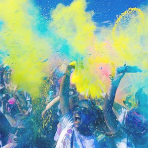 Eine Menschenmenge feiert, indem sie leuchtend blaues und gelbes Pulver in die Luft wirft, wodurch eine lebhafte, bunte Wolke unter einem sonnigen blauen Himmel entsteht. Viele haben ihre Arme erhoben und lächeln oder schreien vor Freude.