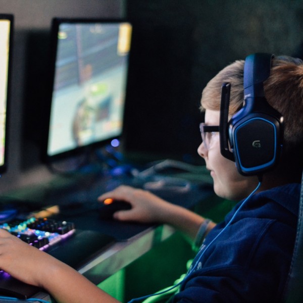Ein Junge mit Brille und Kopfhörern sitzt an einem Schreibtisch und spielt ein Spiel auf einem Computer mit einer Tastatur mit Regenbogenbeleuchtung. Neben ihm steht ein weiterer Monitor, auf dem ebenfalls ein Videospiel zu sehen ist.