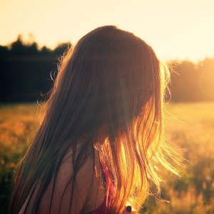 Eine Person mit langem, hellbraunem Haar steht bei Sonnenuntergang auf einer sonnenbeschienenen Wiese und blickt von der Kamera weg. Das warme Sonnenlicht erzeugt einen goldenen Schimmer auf ihrem Haar und dem umliegenden Gras.