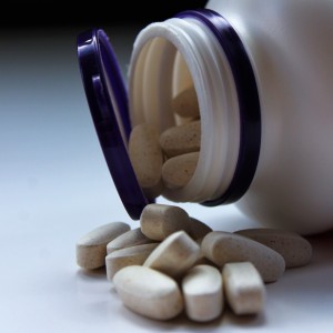 Eine weiße Flasche mit lilafarbenem Deckel liegt auf der Seite und verschüttet mehrere ovale beigefarbene Tabletten auf eine weiße Fläche.