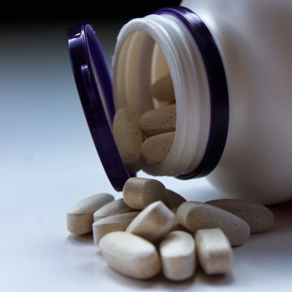 Eine weiße Flasche mit lilafarbenem Deckel liegt auf der Seite und verschüttet mehrere ovale beigefarbene Tabletten auf eine weiße Fläche.