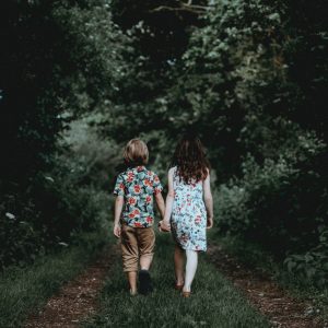 Zwei Kinder, ein Junge und ein Mädchen, gehen Hand in Hand einen schmalen, grasbewachsenen Weg entlang, der von dichtem, grünem Laub und Bäumen umgeben ist. Sie stehen mit dem Rücken zur Kamera und tragen bunte, blumengemusterte Kleidung.