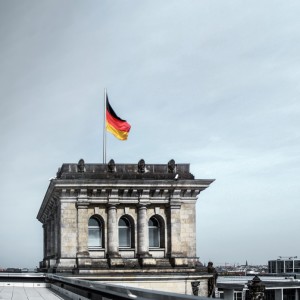 Eine deutsche Flagge weht auf einem historischen Steingebäude, das wahrscheinlich zum Reichstag gehört, unter einem bedeckten Himmel. Im Hintergrund erstreckt sich das Stadtbild von Berlin.