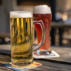 Ein klarer Glaskrug mit hellem Bier und ein Glas dunkles Bier mit Schaum sitzen auf Untersetzern auf einem Holztisch im Freien, mit verschwommenen Menschen und Tischen im sonnenbeschienenen Hintergrund.